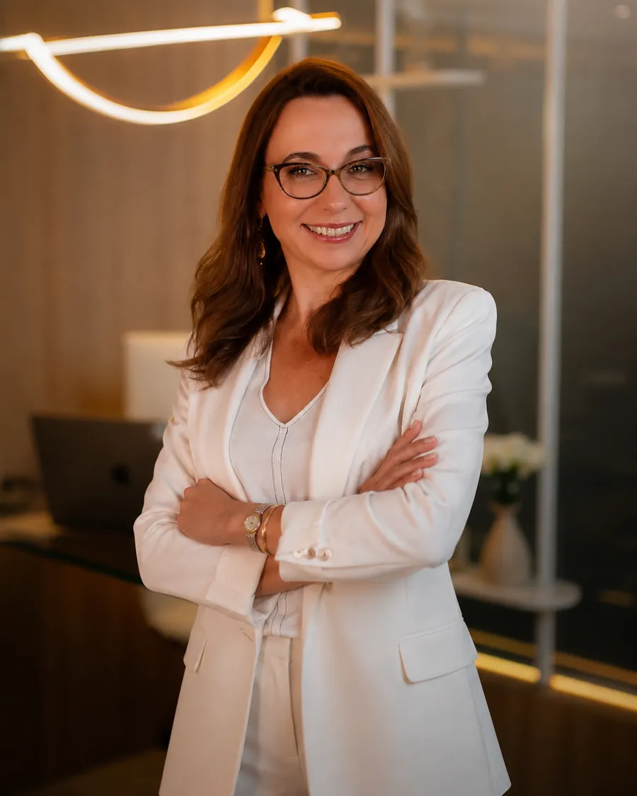 Elaine Cristina de blazer branco, de braços cruzados, no consultório