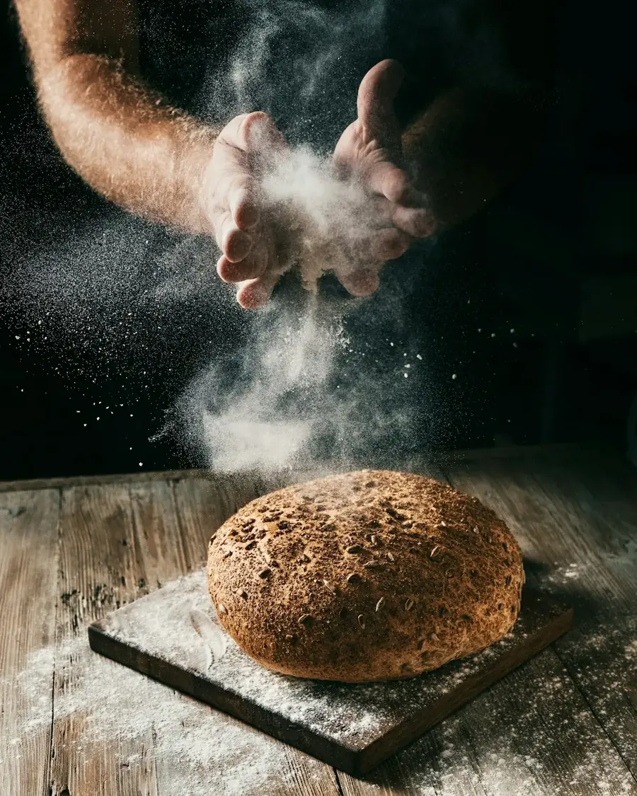 Mãos polvilhando farinha sobre um pão rústico numa bancada de madeira